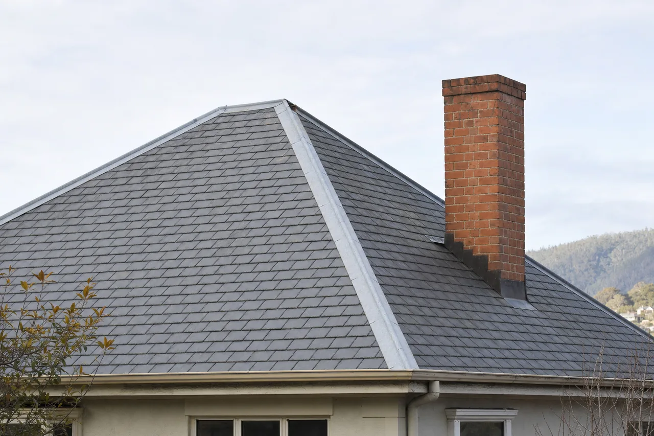 A hipped slate roof with lead-capped hips and a brick chimney against a pale sky — slate roofing, Hobart. Illustrative example.