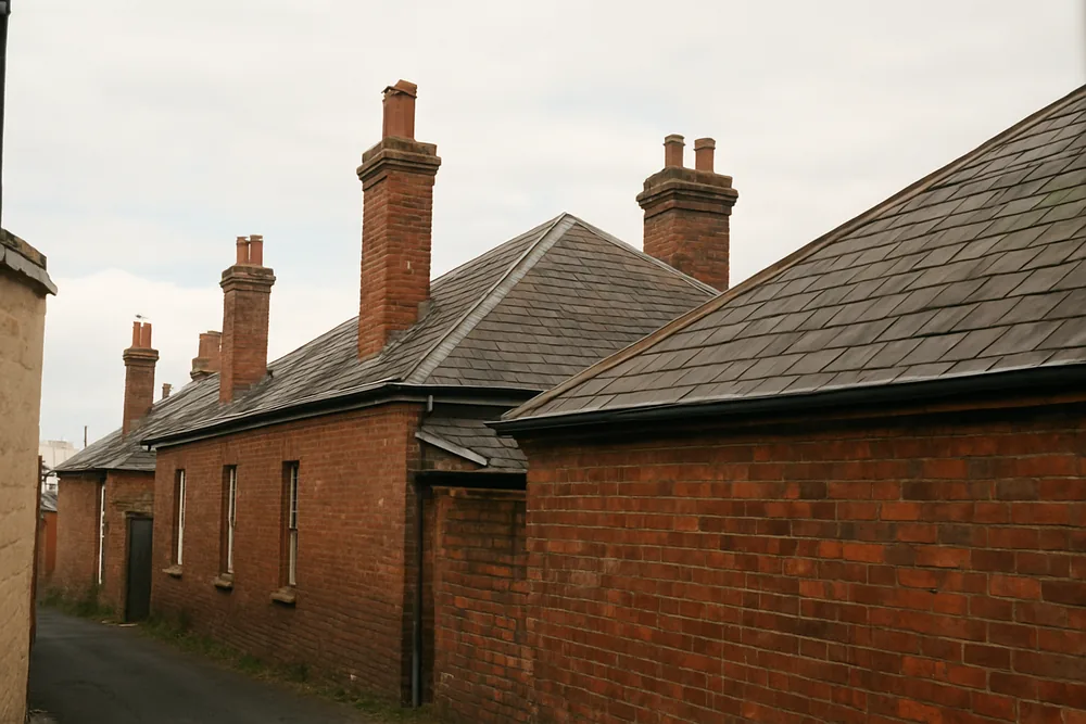 Attached terrace-style roofs in slate with brick chimneys seen from a back lane — slate roofing, Hobart. Illustrative example.